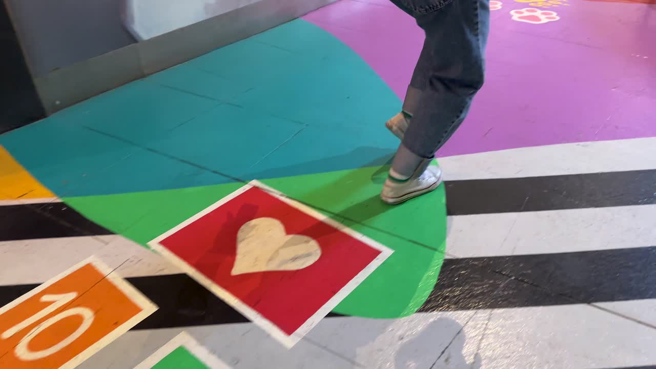 Individual wearing jeans and sneakers hops along a colorful numbered hopscotch path in a brightly lit indoor activity center with patterned flooring