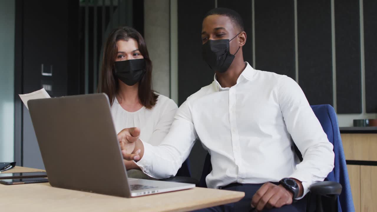 Diverse businessman and businesswoman wearing face masks discussing and using laptop in office