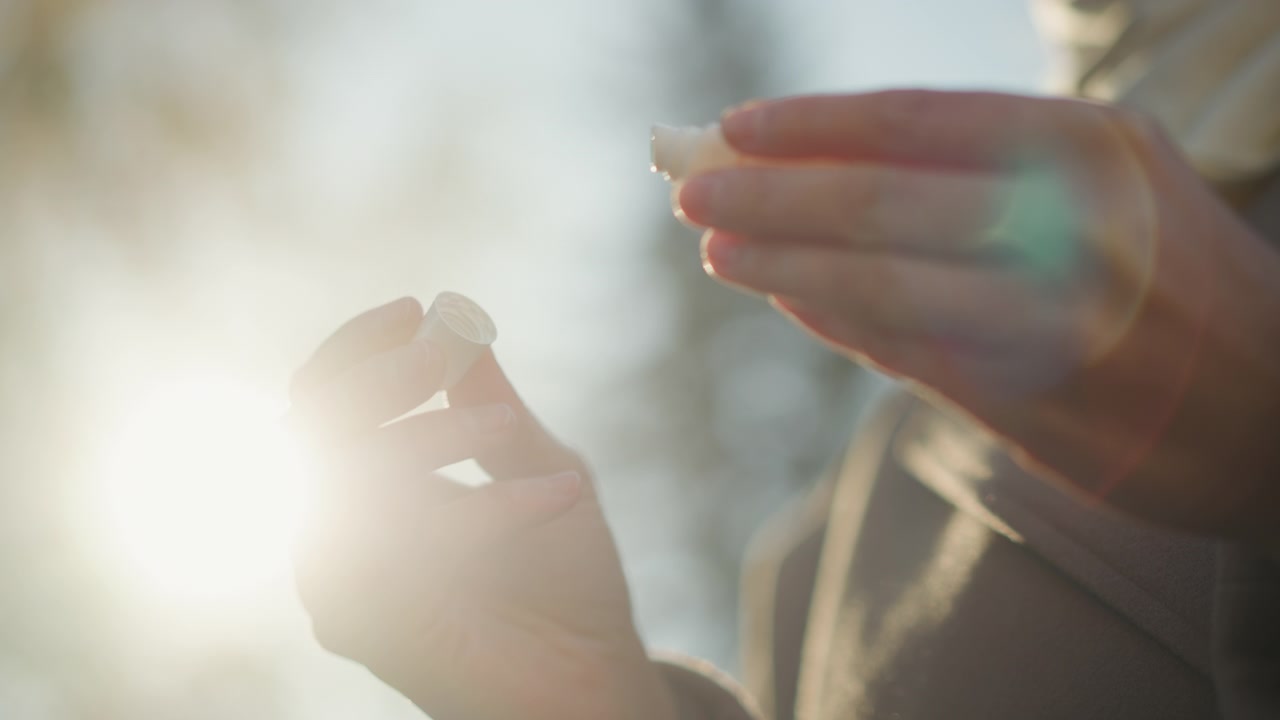 Primo piano delle mani di una persona che tengono e aprono una custodia per lenti a contatto, trasferendo una lente morbida in una mano. La scena presenta una retroilluminazione calda del sole con effetto bokeh che evidenzia la pelle naturale.