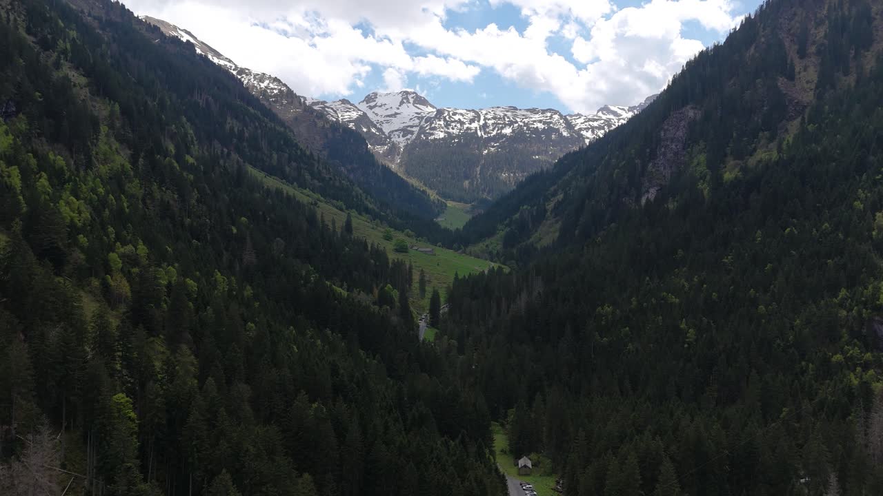 Aerial threatening flight towards a snow-covered mountain in springtime in a valley of the Swiss Alps. It is spring, and the landscape with forests is in bloom. Clouds cover the sky.
