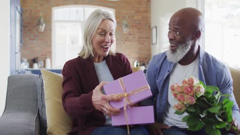 feliz pareja diversa en la sala de estar sentada en el sofá, dando flores y regalos