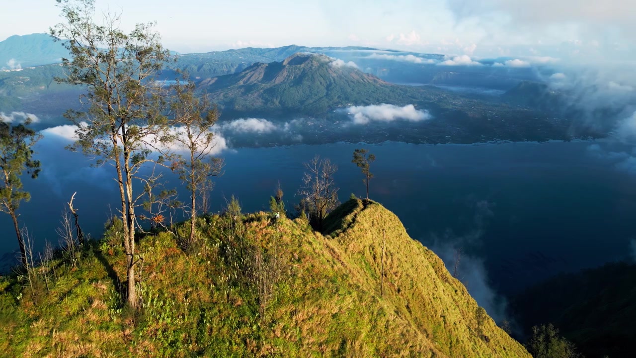 Escursionista solitario che scende un sentiero stretto su una cresta su un ripido pendio di montagna sopra un lago vulcanico con paesaggio vulcanico e nuvole basse che si muovono ad alta quota in pieno giorno, ripresa aerea statica