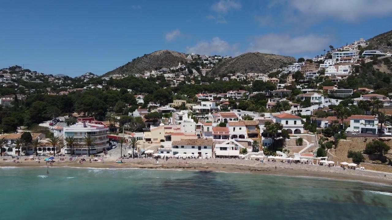Voo ascendente de drone revela uma vista aérea deslumbrante da praia de El Portet em Moraira, Espanha, exibindo águas cristalinas azul-turquesa e uma costa acidentada