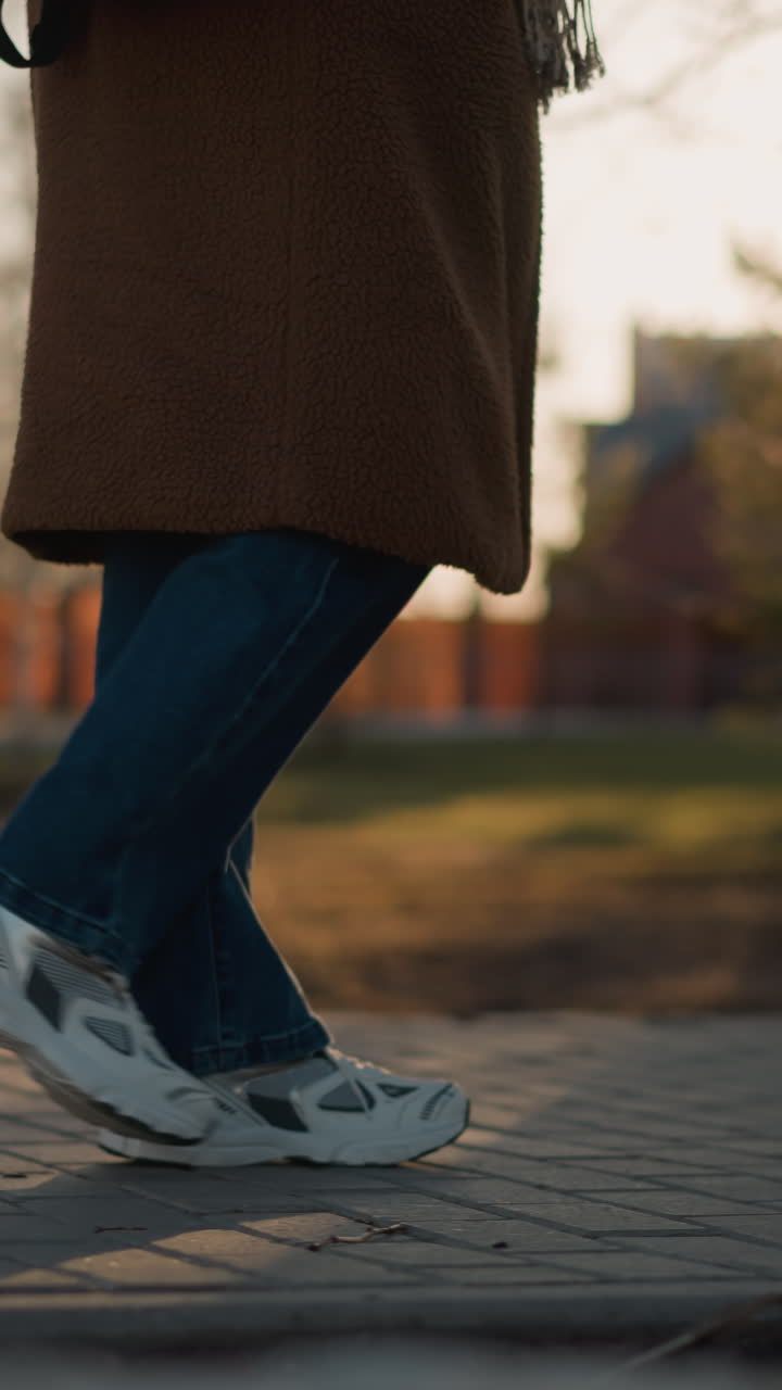 A person wearing a brown coat, jeans, and white shoes is shown walking slowly through a park at sunset. The focus is on their legs, emphasizing their slow, sorrowful steps