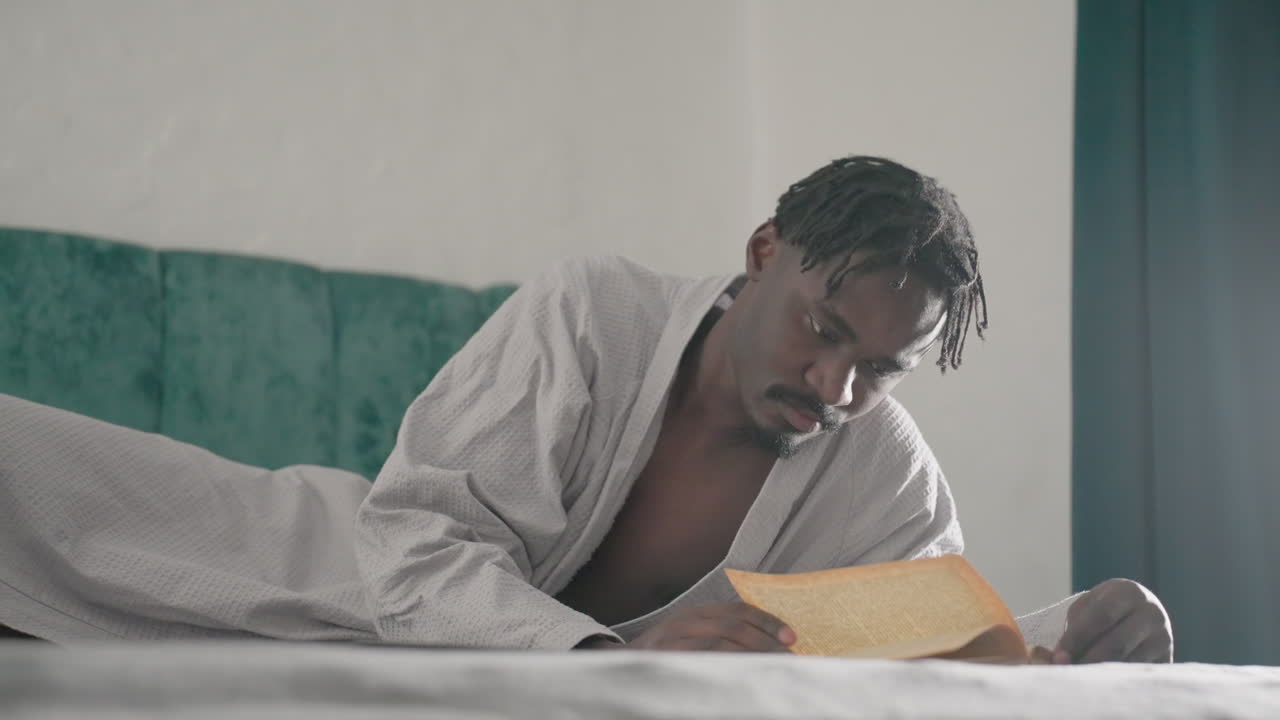 Man in white robe lying on bed, deeply focused on reading large book, slowly turning pages with intense concentration. Soft natural light from window creates calm and reflective indoor atmosphere