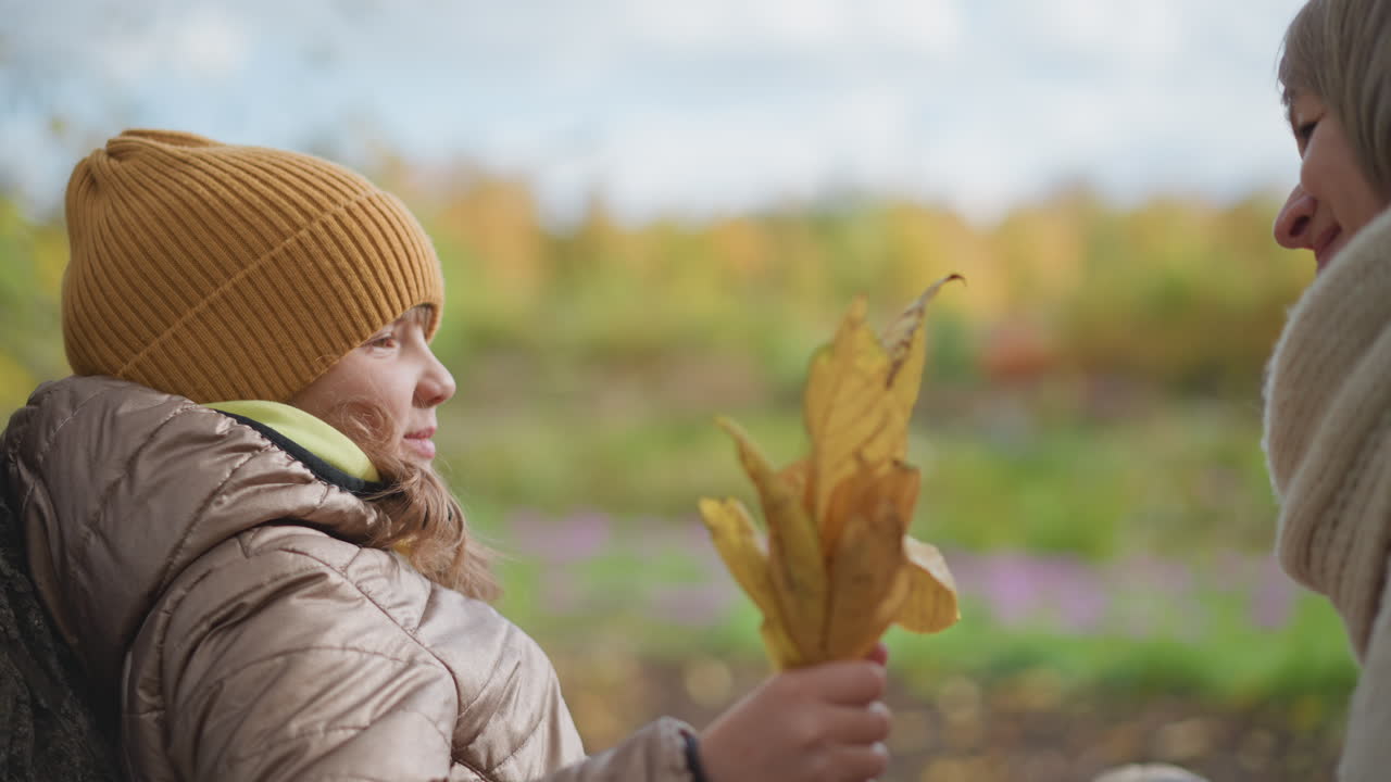 young girl rests quietly on tree trunk wearing cozy jacket and mustard beanie while mother offers bundle of dry autumn leaves and gives her a tender warm hug in colorful serene fall park setting