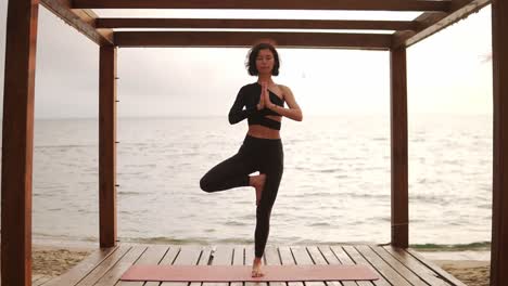 Retrato-De-Una-Mujer-Con-Ropa-Deportiva-Negra-Practicando-Yoga-En-Una-Alfombra-Frente-Al-Mar-Por-La-Mañana-Realizando-Una-Pose-De-Una-Pierna