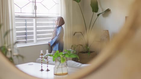 video de una mujer biracial pensativa en hijab mirando por la ventana en la sala de estar reflejada en el espejo