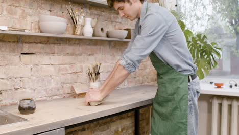 clerk in agreen apron kneading clay on top of a table in a pottery workshop