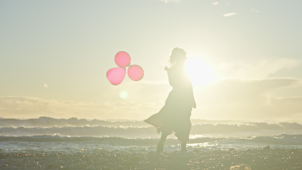 젊은 여성은 해가 그녀의 뒤에 지면 세 개의 빨간 풍선을 들고 해변을 따라 걷는다