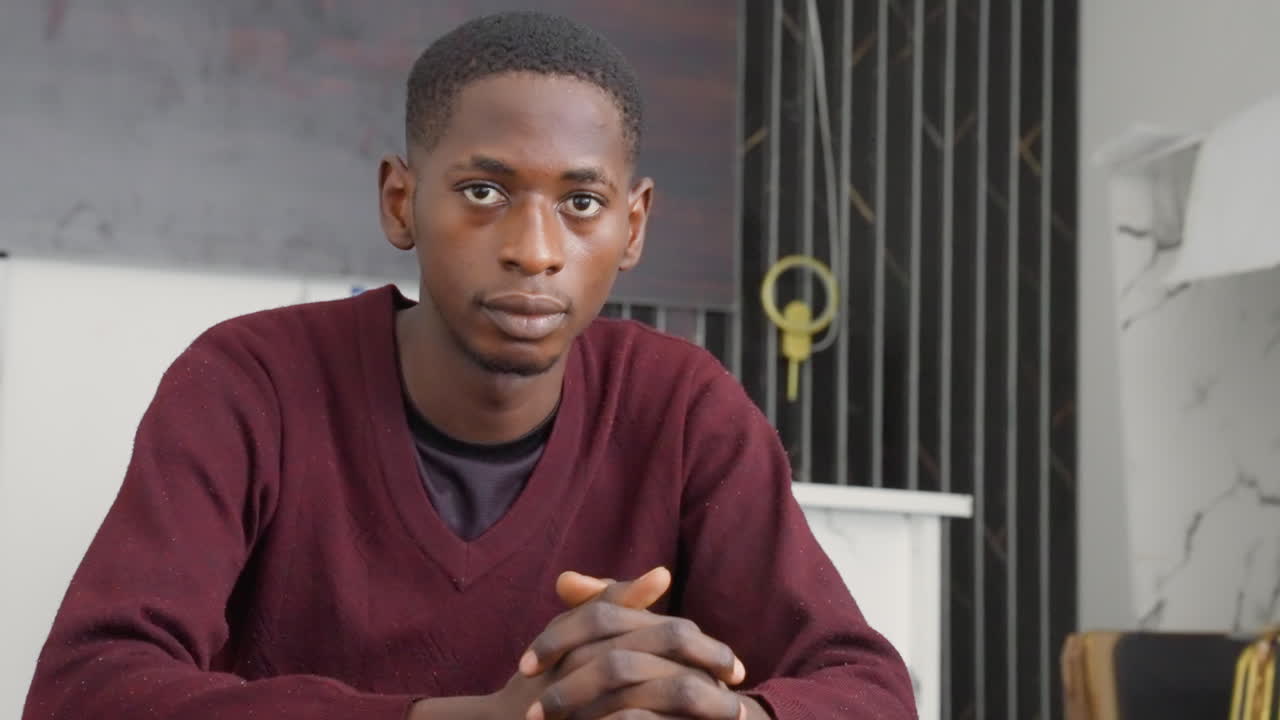 Uomo di colore contemplativo davanti a un quadro nero, mani giunte sulla scrivania, maglione bordeaux, interno moderno con arredamento minimalista, sguardo pensieroso che suggerisce uno scrittore o un consulente che si prepara per un'intervista.
