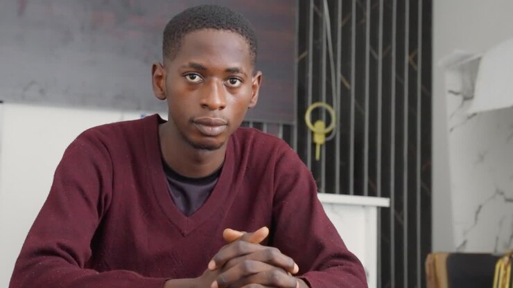 Contemplative black man before black painting, hands clasped on desk, maroon sweater, modern interior with minimalist decor, thoughtful gaze suggesting writer or consultant preparing for interview
