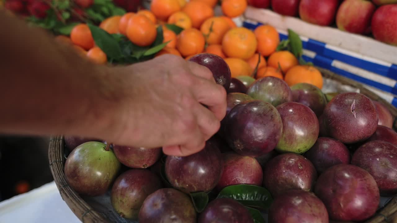 Étale de marché de rue au Vietnam avec un vendeur sélectionnant à la main des prunes mûres dans un panier tressé, des mandarines et des pommes en arrière-plan, texture en gros plan, ambiance de produits frais et lumière matinale