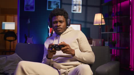 young man sitting on sofa at home relaxing by playing videogames on gaming console, unwinding in neon lit apartment by competing in online multiplayer e sports tournament. camera b.