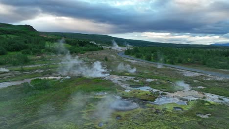 aerial view of steaming vents and bubbling geothermal pools in the geysir geothermal field, surrounded by lush greenery and dramatic skies.