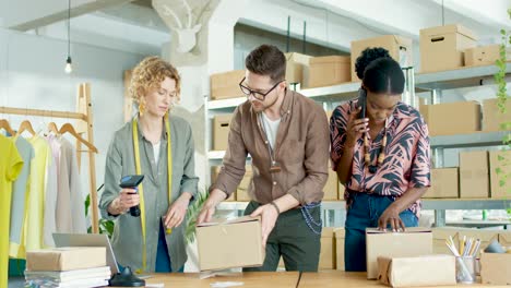 multiethnic designers team wrapping parcels and closing boxes with sticky tapes while african american woman manager talking on smartphone with a client