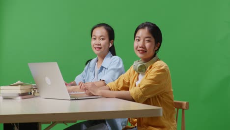 asian woman students writing and typing on a laptop then smiling to camera while sitting on a table in the green screen background classroom