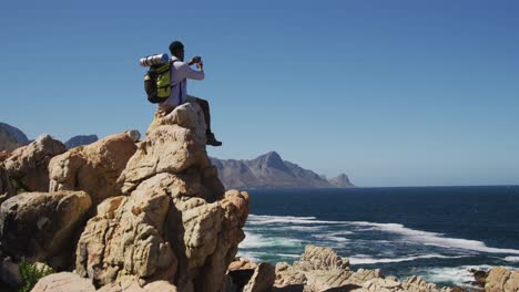 african american man hiking taking pictures with smartphone sitting on rock by the coast