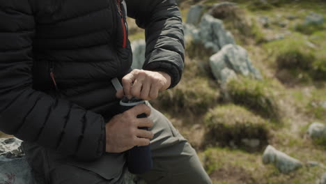 Close-up-shot-of-mans-lap-unscrewing-the-top-of-his-drinking-bottle,-sitting-on-a-rock-in-the-mountains