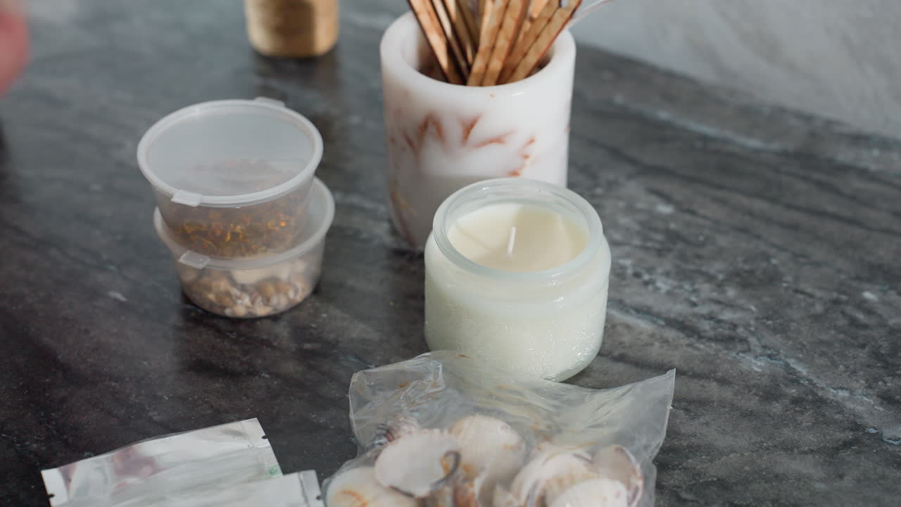 Close-up of person dropping shopping items including sealed food packet and seashells on marble table near lit white candle and container of wooden sticks