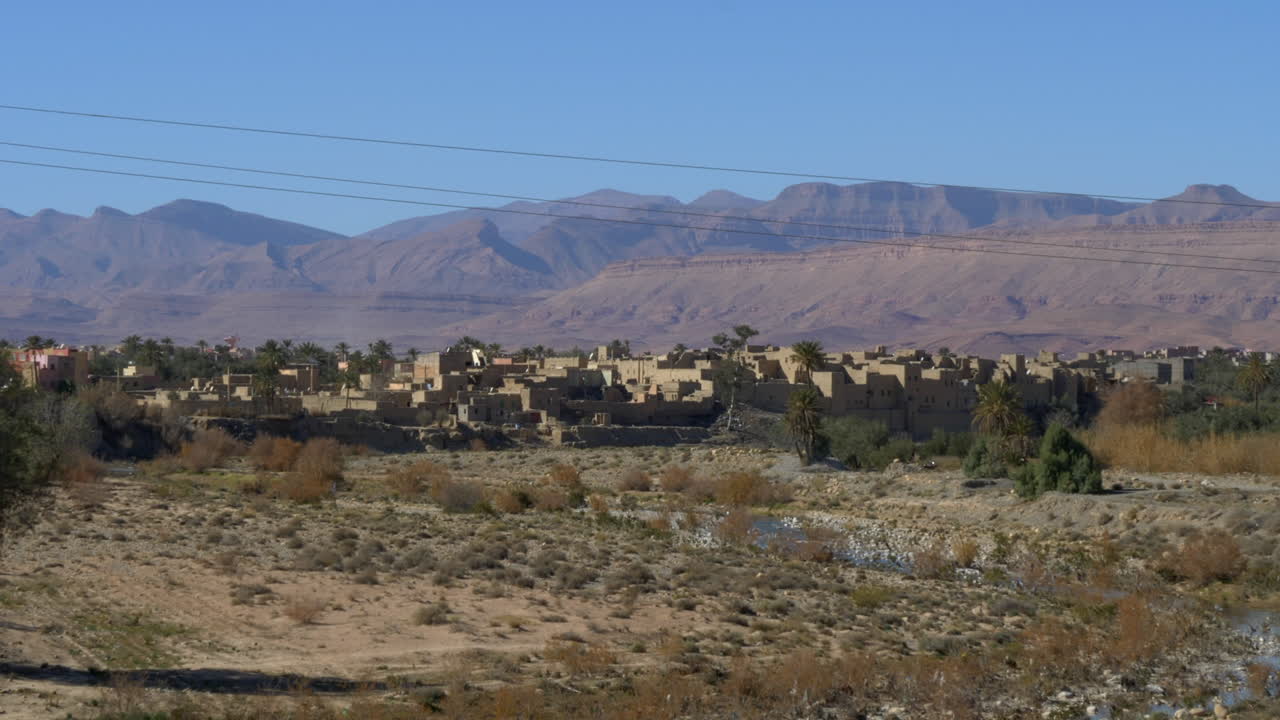 toma panorámica lenta de un pueblo del desierto marroquí con un telón de fondo de altas montañas en un día soleado