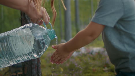 woman is pouring water from big bottle on hands of little boy child is washing hands at forest camp