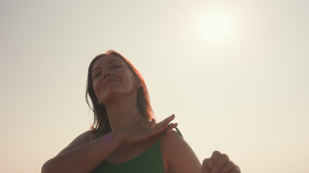 young woman in green dress stands in sunlit grassy field with eyes closed, hands behind head gently scratching hair, breathing fresh air, enjoying warm breeze and golden scenery