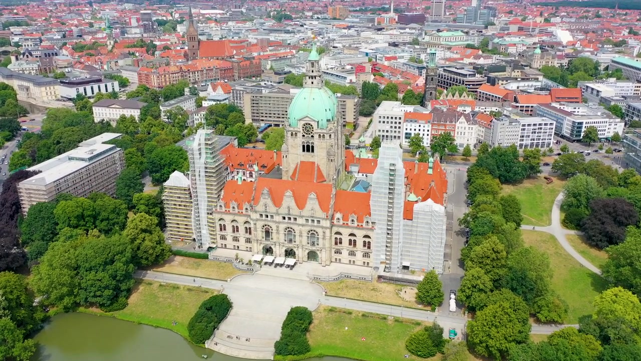 Cinematic aerial shot showcasing the iconic New City Hall in Hanover, Germany, surrounded by green trees and a tranquil pond, perfect for travel, urban, or historical projects.