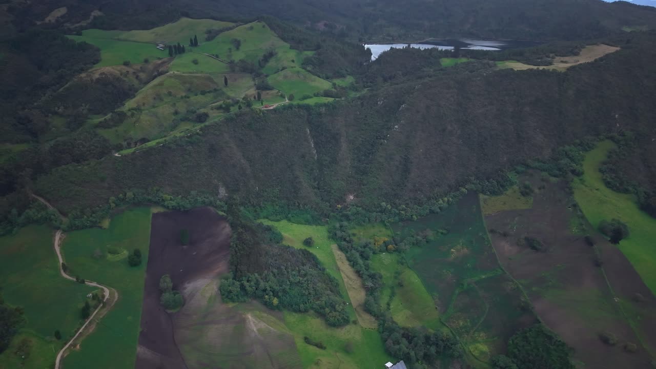 A drone panning shot over volcanic slopes and green fields near Zipaquirá, revealing the distant Laguna de Pantano Redondo under soft daylight