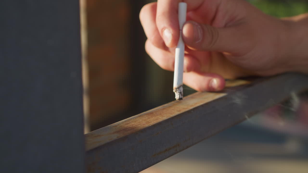 close up of fair skinned person's hand holding cigarette and pressing burning tip against metal surface while smoke rises gently in outdoor environment under warm daylight