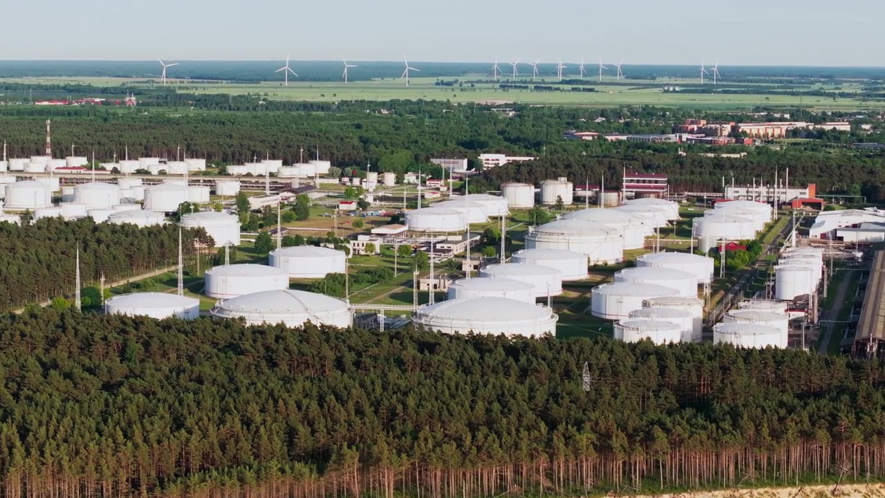 Telephoto drone footage overlooking a large oil storage terminal with white fuel tanks and wind turbines in the distance near Ventspils, Latvia. Aerial view highlighting industrial infrastructure