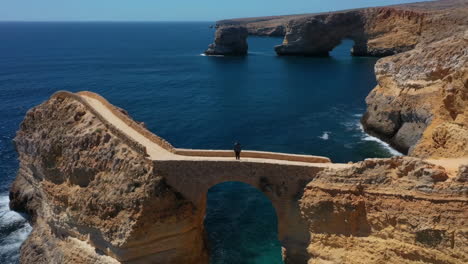 person standing on a stone arch bridge over the ocean with natural rock formations