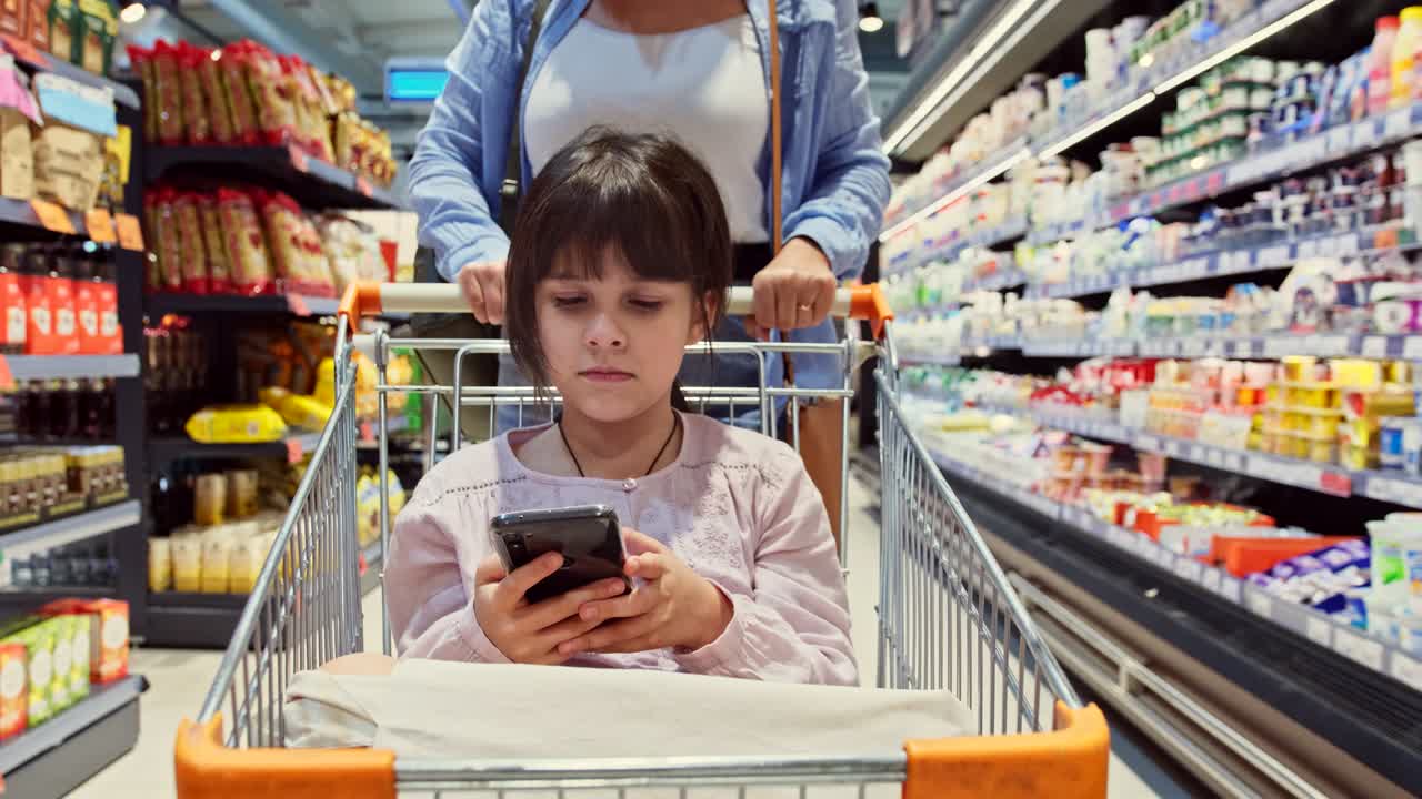 식료품 바구니에서 스마트폰을 들고 있는 소녀의 클로즈업 사진과 뒤에서 매장의 선반 사이에서 바구니를 밀고 있는 어머니의 사진.