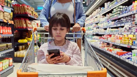 foto en primer plano de una niña en el carrito de comestibles mirando el teléfono inteligente en sus manos y su madre en el fondo empujando el carrito entre los estantes de la tienda..