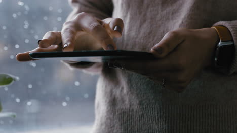 cerca de las manos de la mujer usando una tableta digital navegando por mensajes en línea leyendo redes sociales disfrutando de un dispositivo móvil con pantalla táctil de pie junto a la ventana relajándose en casa en un día frío y lluvioso