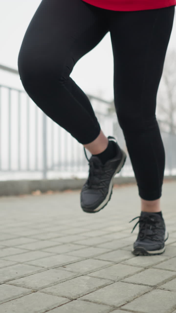 lower angle view of athlete in black sneakers and leggings jogging along iron railing of bridge in foggy winter atmosphere with faint cityscape and bare trees in background showing cold fitness