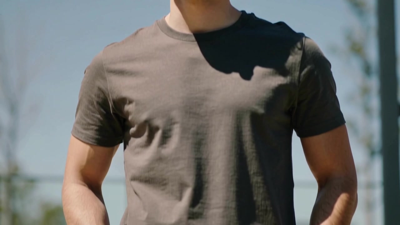Hombre sonriente buscando calma, la cámara baja mientras él mete las manos en los bolsillos de sus jeans junto a un poste en la plaza, vistiendo una camiseta.