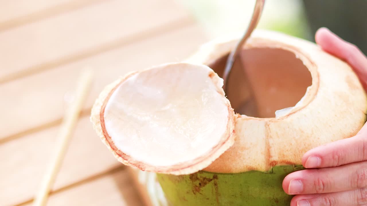 Hands holding a fresh coconut with a spoon, under bright daylight, capturing a tropical refreshment moment
