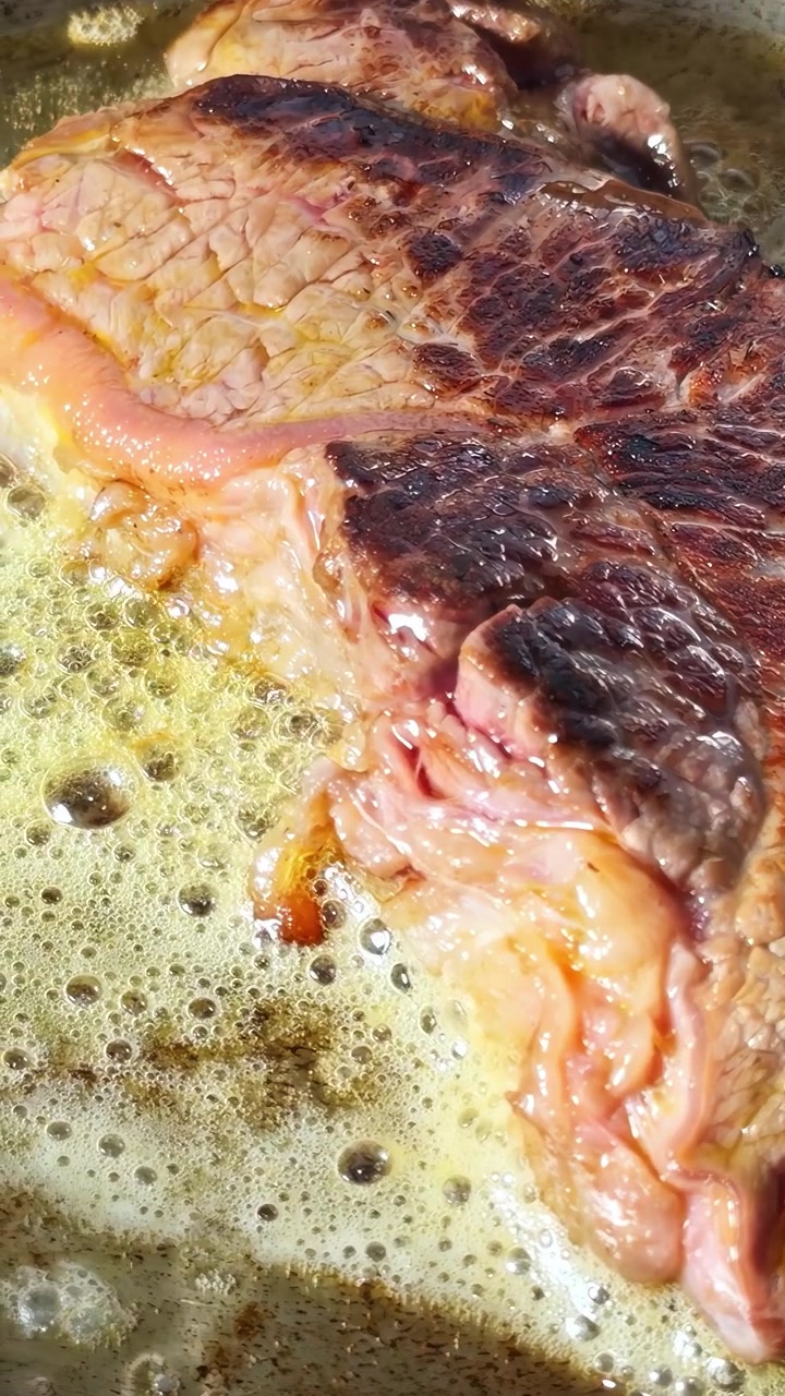 Una succulenta bistecca sfrigola in padella, la sua superficie si caramella nell'olio caldo. Perfetto per video culinari o tutorial di cucina.