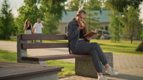 lady seated on park bench eating burger while reading outdoors, with two people walking in the background and a relaxed park setting, with trees, benches, and a sunny atmosphere