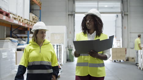 trabajadoras multiétnicas en casco caminando y contando bienes en stock