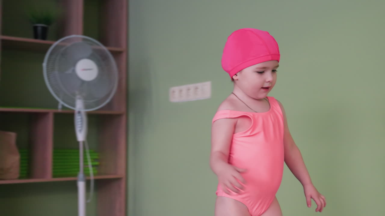 Toddler bends to pick tennis ball from shelf, small hand reaching down, focused gaze, soft indoor light, neat storage rack visible, careful posture, playful learning moment, calm poolside area
