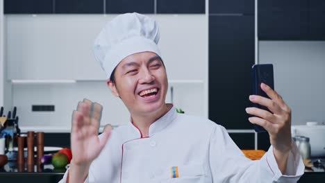 집 주방에 서 있는 동안 스마트폰으로 비디오 통화를 하는 아시아인 셰프의 손을 흔들고 있습니다.