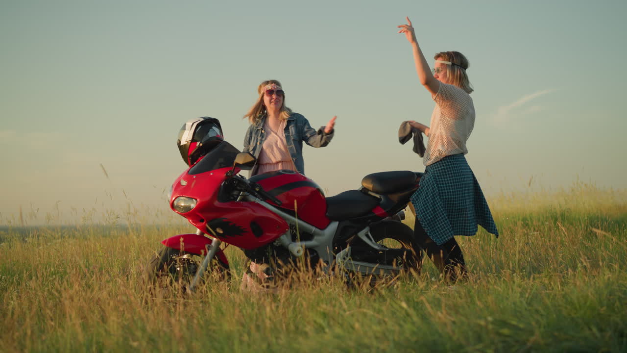 두 명의 즐거운 여성이 은 하늘 아래 잔디에 있는 빨간 모터사이클 근처에서 춤을 추고 있으며, 그들 중 한 명은 그들이 축하하고 야외에서 시간을 즐기면서 청소 천을 들고 있습니다.