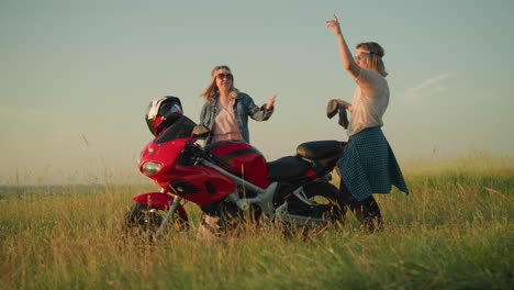 two joyful women are dancing near a red motorcycle in a grassy field under the clear sky, one of them is holding a cleaning cloth as they celebrate and enjoy their time outdoors