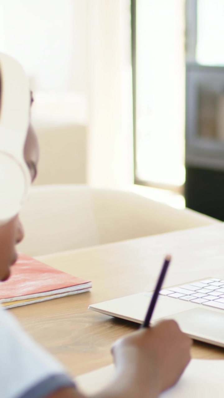 Vertical video: Panning causing child with headset writing homework at home table with laptop marks