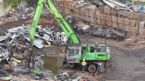 drone captures a green excavator sorting metal scraps in a gold coast junkyard. overcast lighting highlights industrial activity
