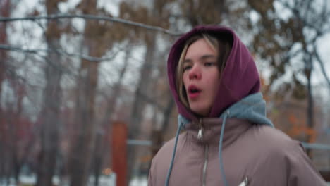 young lady in winter outfit jogging because of cold in outdoor park with trees and snow, urban setting with playground in background, breath visible due to cold weather