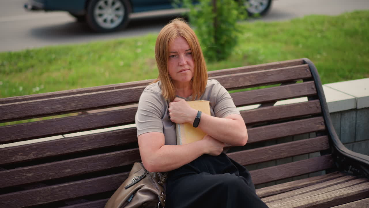 Woman sitting alone on bench outdoors reading book, flipping to new page then closing it and pressing to chest in thoughtful emotion and blurred street background with pigeons feeding nearby