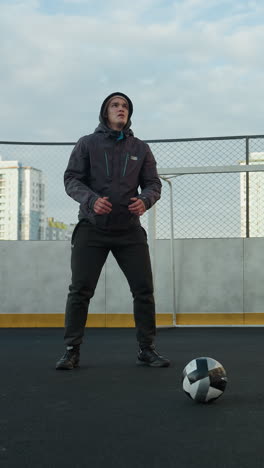deportista realizando sentadillas con las manos juntas en un campo de deportes al aire libre preparándose para el ejercicio, una pelota de fútbol en primer plano, edificios urbanos de gran altura y postes de gol